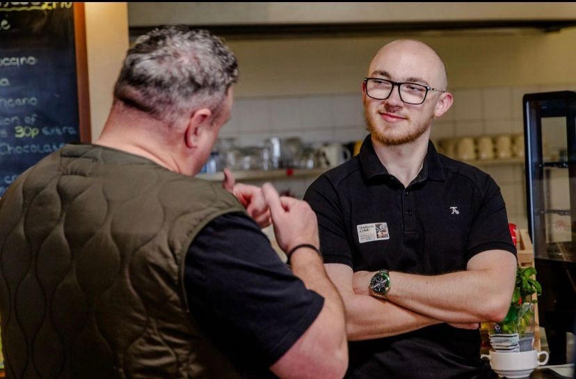 Two people in a cafe having a conversation, one wearing glasses and a name tag.