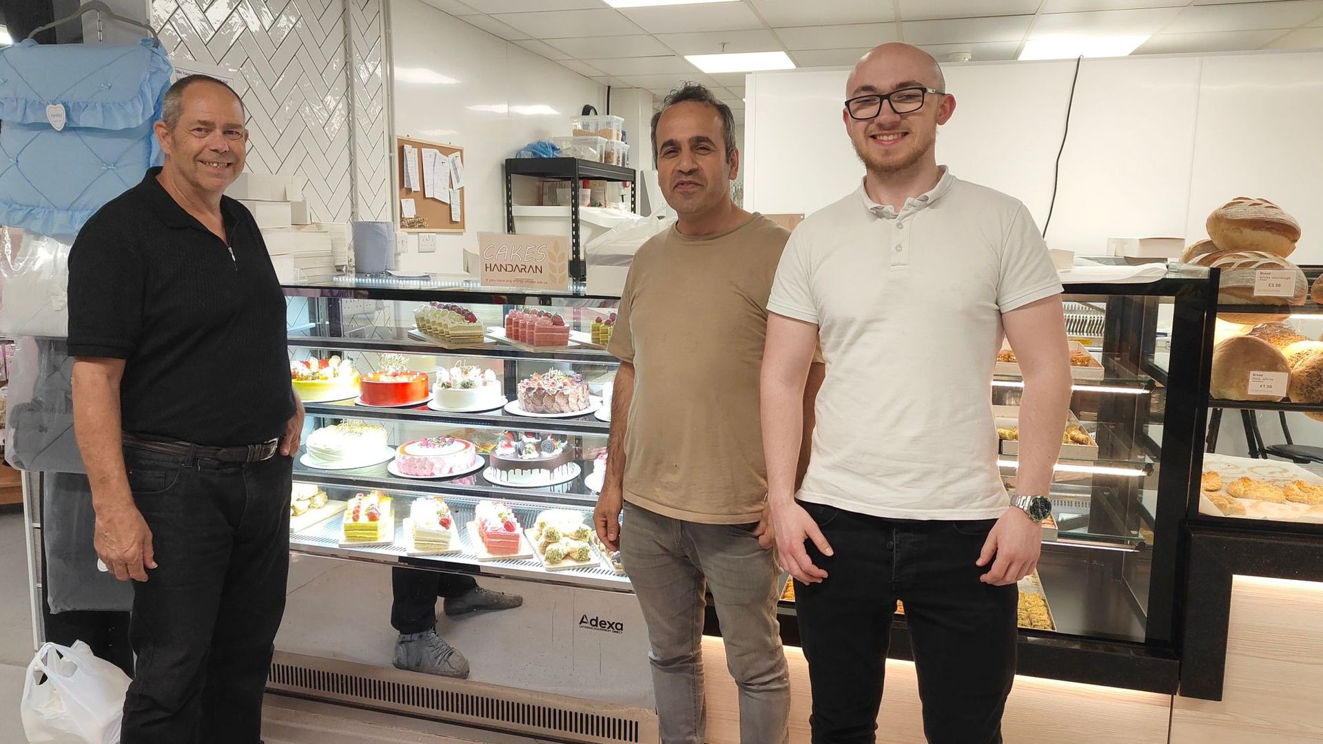 Three men standing in front of a bakery display case filled with various cakes and pastries.