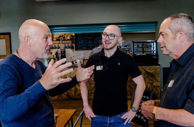 Three men in conversation inside a café, with a chalkboard menu and counter in the background.